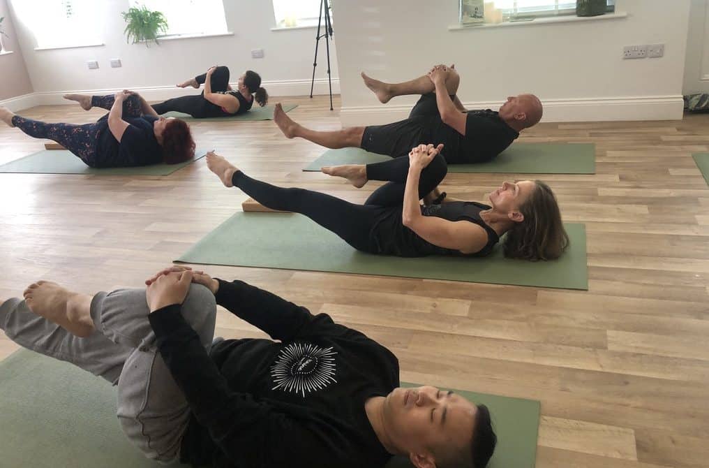 men and women laying on a yoga mat on the floor of Shine With Tabitha Yoga Studio with their legs in the air practicing Pilates