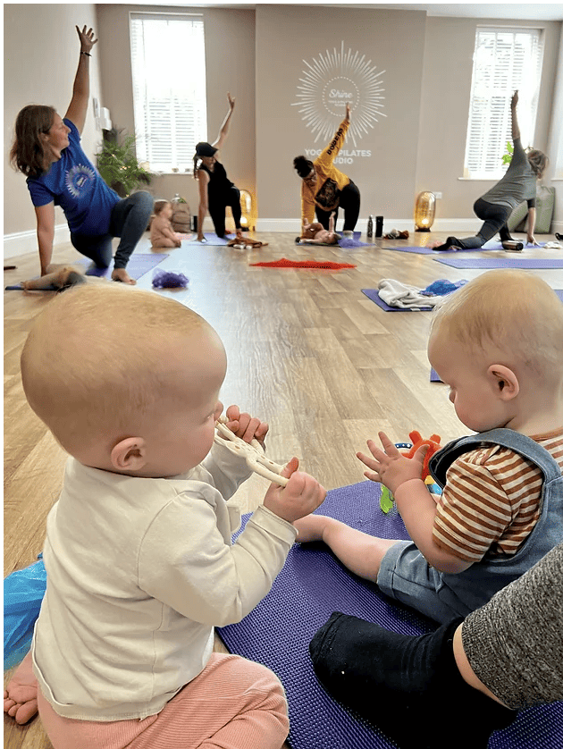 2 babies sitting on the floor playing while mothers and other babies are practising yoga at Shine With Tabitha Yoga